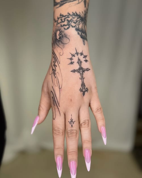 A delicate black and grey ornate cross tattoo on the hand with fine line script and minimal design — by otaybeatdown, los angeles