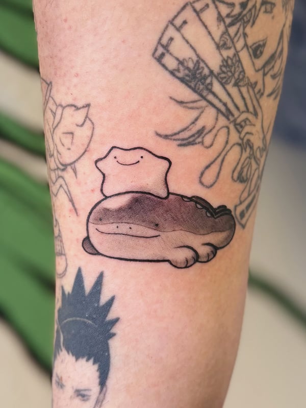 A cute anime-style tattoo of a smiling blob-like creature sitting on a larger creature, with simple linework and a mix of black and grey shading on the arm — by Tweetwee, west covina, ca