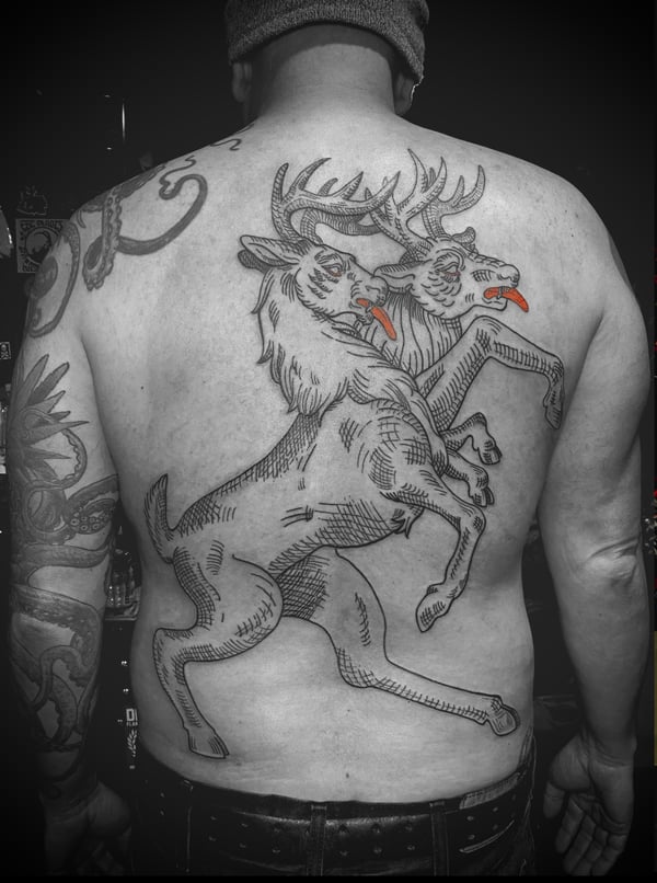 An illustrative blackwork tattoo of two stylized deer with red tongues on the back, featuring bold lines and minimal color accents — by Tag Devilish, wrightwood, ca
