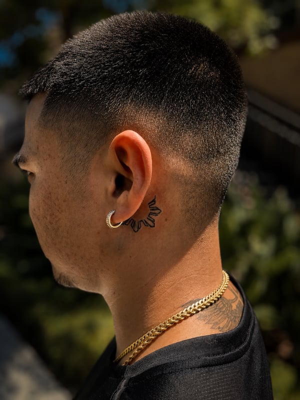 A minimalist black line sun tattoo behind the ear, featuring simple linework — by HEAVY INK, chino, ca