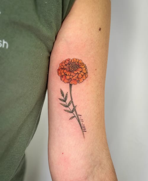 A delicate marigold flower tattoo in fine line style on the inner arm, featuring orange petals and a date in script — by Melanie Benoit, los angeles