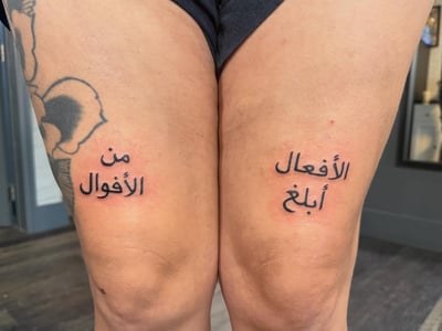 A minimalist black Arabic script tattoo on the thighs, featuring a quote in simple lettering — by Kiera Garvin, los angeles