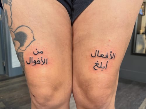 A minimalist black Arabic script tattoo on the thighs, featuring a quote in simple lettering — by Kiera Garvin, los angeles