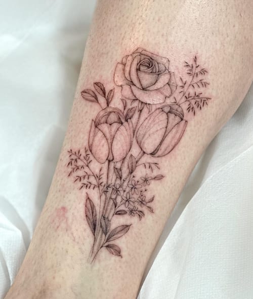A delicate black and grey floral bouquet tattoo on the leg featuring roses and tulips in fine line style — by Melanie Benoit, los angeles