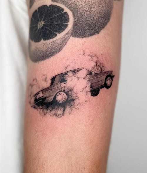A black and grey illustrative tattoo of a car on the forearm, featuring dynamic shading and a smoky effect — by Level tattoo, los angeles
