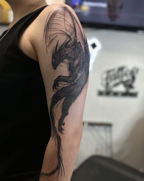 A bold black and grey sketch style dragon tattoo on the upper arm — by Maddie Ramirez, los angeles