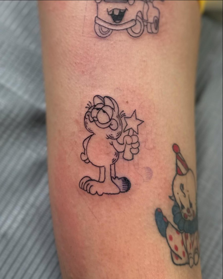 A black and grey minimalist linework tattoo of Garfield holding a star on the forearm — by Maddie Ramirez, los angeles