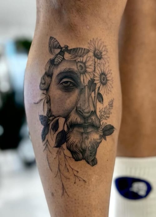 A black and grey tattoo on the calf depicting a realistic face intertwined with flowers and leaves, showcasing intricate detail and shading — by otaybeatdown, los angeles