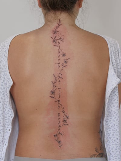 A delicate black and grey script tattoo with flowers running along the spine — by Melanie Benoit, los angeles
