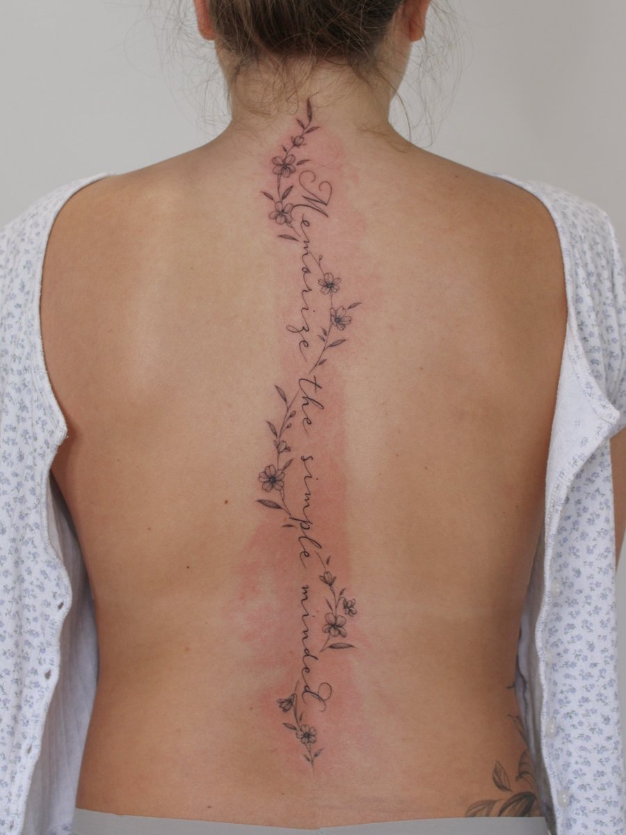A delicate black and grey script tattoo with flowers running along the spine — by Melanie Benoit, los angeles