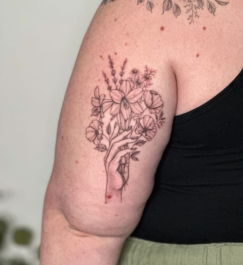 A delicate fine line tattoo of a hand holding a bouquet of flowers on the upper arm, done in black and grey — by Melanie Benoit, los angeles