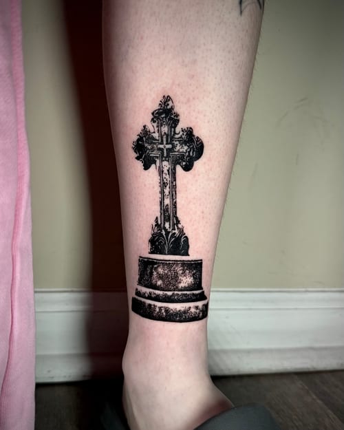 A black and grey realistic depiction of an ornate cross on the calf with detailed shading and texture — by emily, los angeles