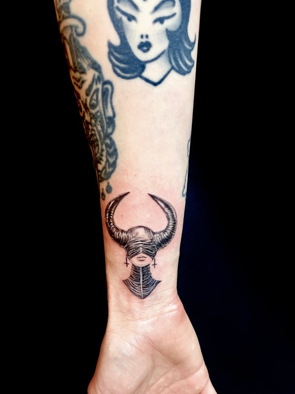 A black and grey linework tattoo of a woman with horns on the forearm, featuring bold outlines and minimal shading — by zitronxus, los angeles