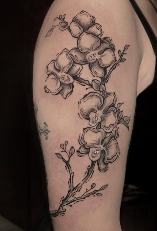 A black and grey fine line floral tattoo on the upper arm, featuring delicate flowers and branches with intricate linework — by Shelby Bianco, los angeles