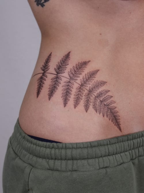 A delicate black and grey fern leaf tattoo on the rib, featuring fine line botanical details — by Melanie Benoit, los angeles