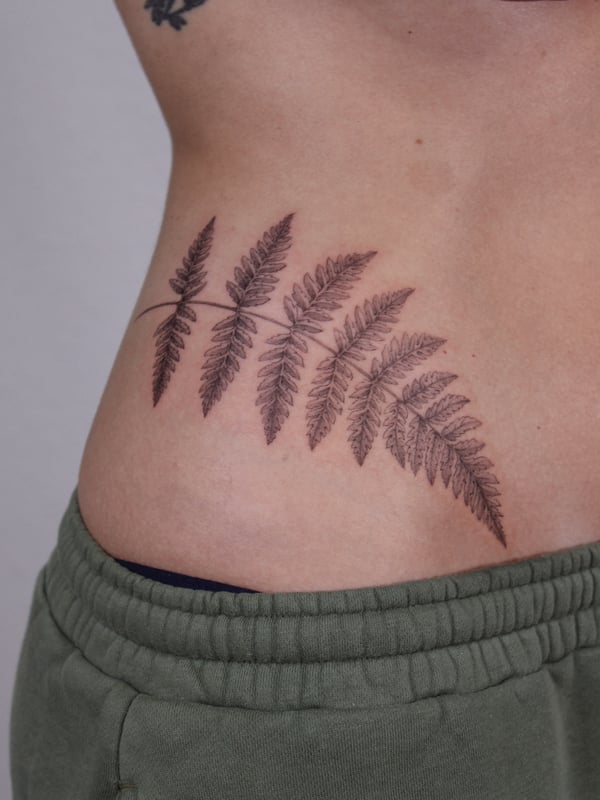A delicate black and grey fern leaf tattoo on the rib, featuring fine line botanical details — by Melanie Benoit, los angeles