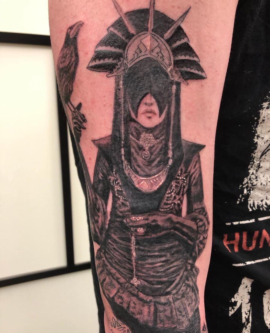 A dark and detailed black and grey tattoo of a mysterious figure with a raven on the forearm, featuring ornate headgear and intricate shading — by Sandra Price, los angeles