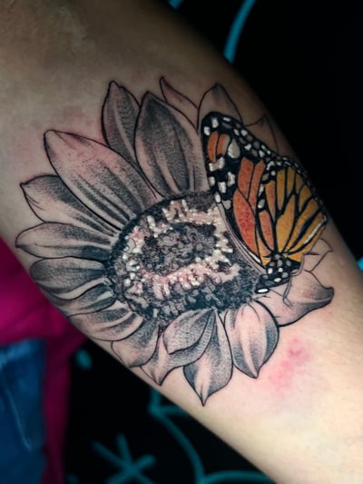 A realistic tattoo of a butterfly on a black and grey flower, located on the arm. The butterfly is in color, contrasting with the monochrome flower — by Josue Bravo, los angeles