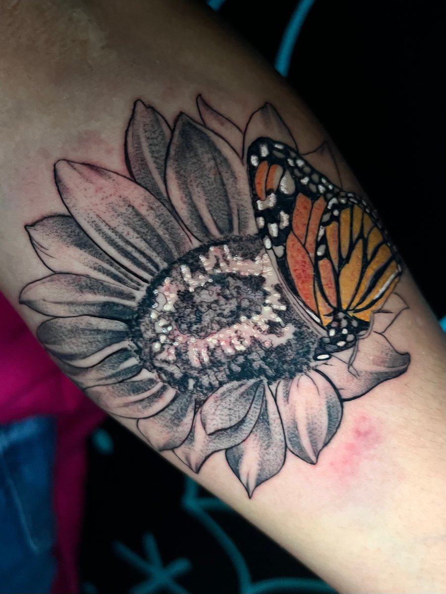 A realistic tattoo of a butterfly on a black and grey flower, located on the arm. The butterfly is in color, contrasting with the monochrome flower — by Josue Bravo, los angeles