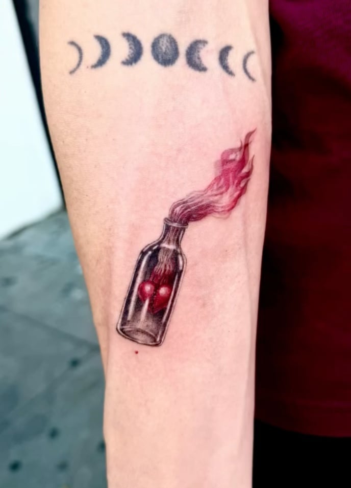 A small tattoo on the forearm featuring a realistic bottle with red liquid and smoke, accompanied by a series of moon phases in black ink above it — by zitronxus, los angeles