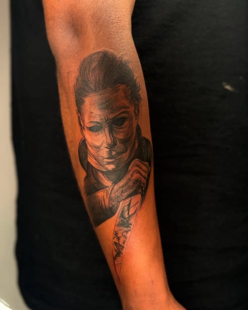 A realistic black and grey tattoo of a masked figure holding a knife on the forearm, with a dark and graphic style — by zealous, los angeles