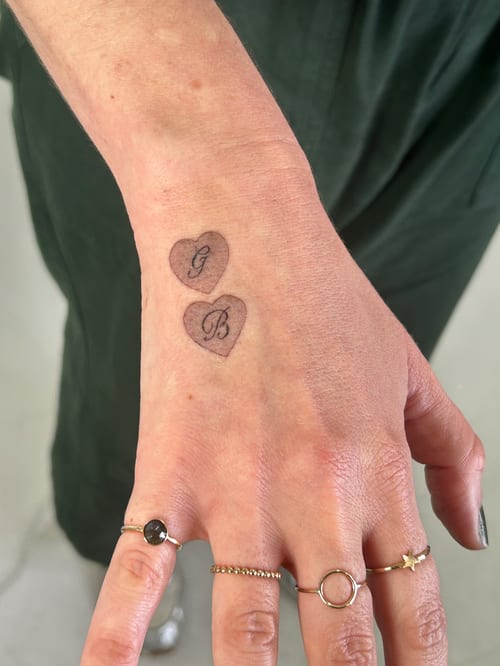 A minimalist black and grey tattoo of two hearts with initials 'G' and 'B' on the hand — by Brian Kies, los angeles