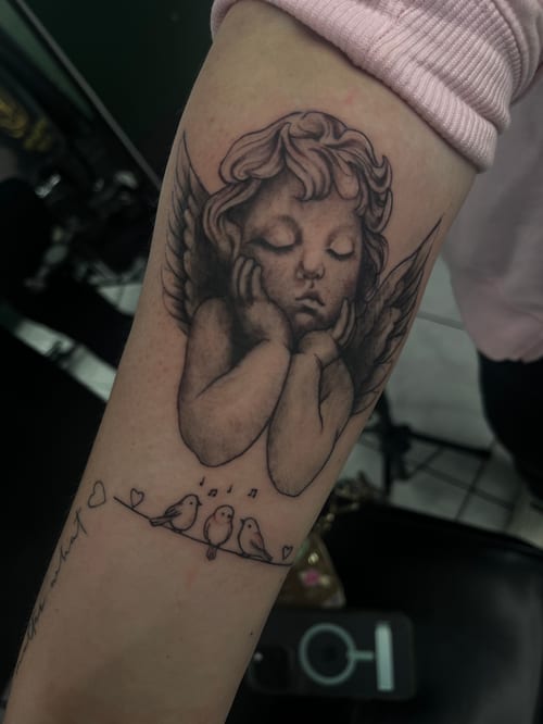A black and grey tattoo of a sleeping angel with wings on the forearm, accompanied by three small birds on a branch with musical notes — by Mr._Folks, los angeles