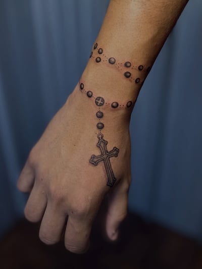A delicate black and grey rosary tattoo on the wrist with a cross extending onto the hand, featuring dotwork detailing — by Maddie Ramirez, los angeles