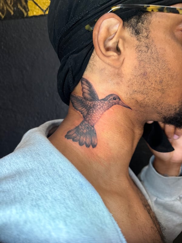 A realistic black and grey hummingbird tattoo on the neck, showcasing delicate details and shading — by Kiera Garvin, los angeles