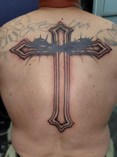 A large black and grey cross tattoo on the back with a crown of thorns, featuring bold outlines and shading — by anoetic.designs.ultd, los angeles