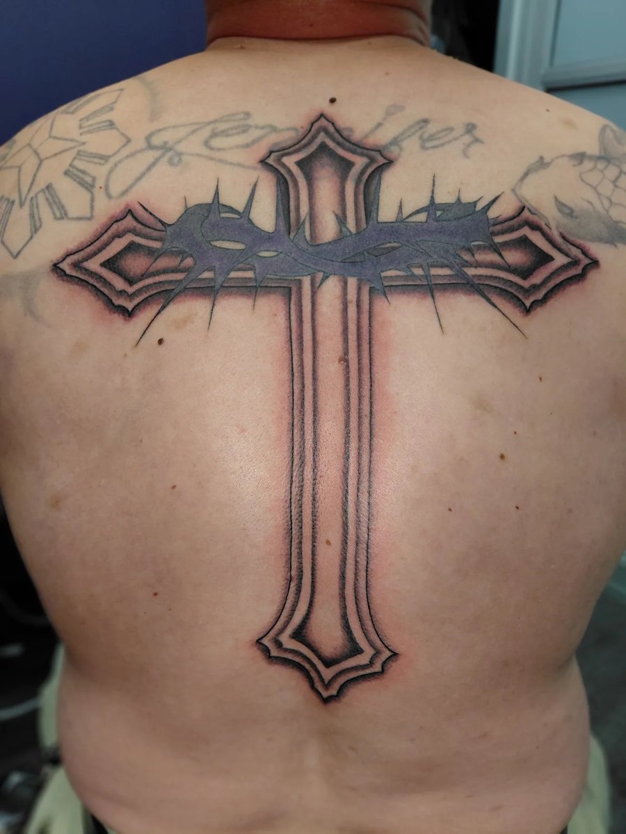 A large black and grey cross tattoo on the back with a crown of thorns, featuring bold outlines and shading — by anoetic.designs.ultd, los angeles