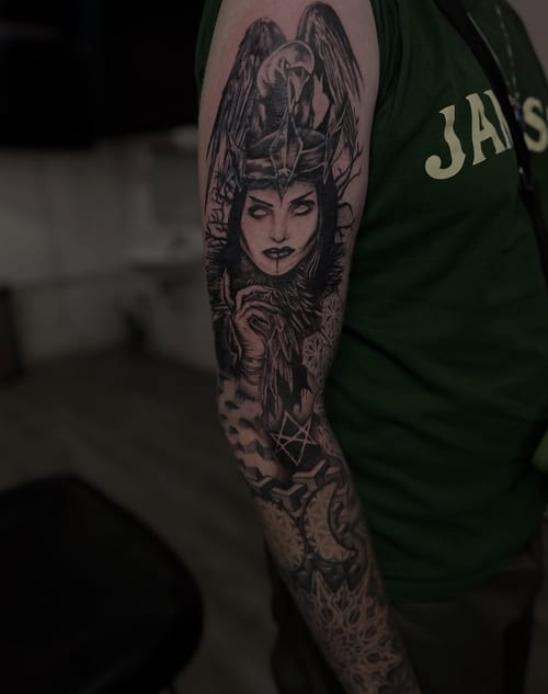 A detailed black and grey full sleeve tattoo featuring the Morrigan goddess with a raven, showcasing intricate shading and mythological elements — by Maddie Ramirez, los angeles