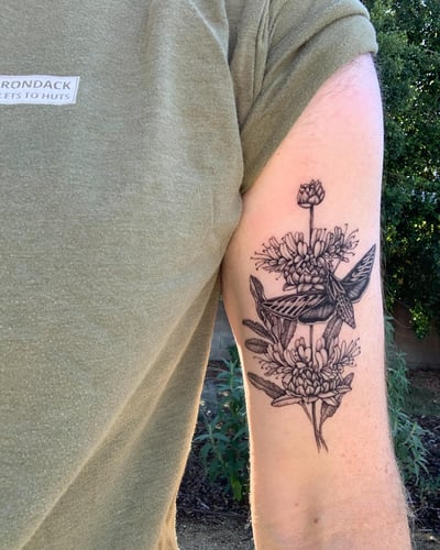 A detailed black and grey tattoo of a moth and flowers on the upper arm, featuring intricate linework and botanical elements — by Melanie Benoit, los angeles
