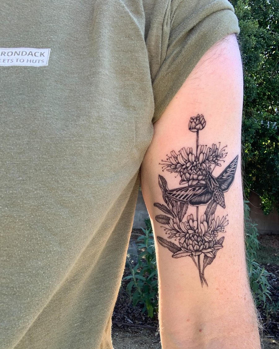 A detailed black and grey tattoo of a moth and flowers on the upper arm, featuring intricate linework and botanical elements — by Melanie Benoit, los angeles