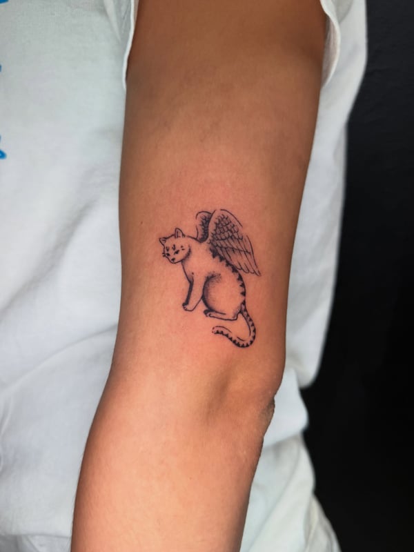 A minimalist black and grey tattoo of a cat with wings on the forearm, featuring delicate linework — by Kiera Garvin, los angeles