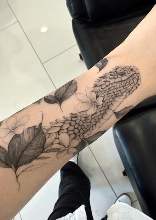 A black and grey fine line tattoo of a snake intertwined with flowers on the forearm, featuring detailed scales and delicate floral elements — by otaybeatdown, los angeles