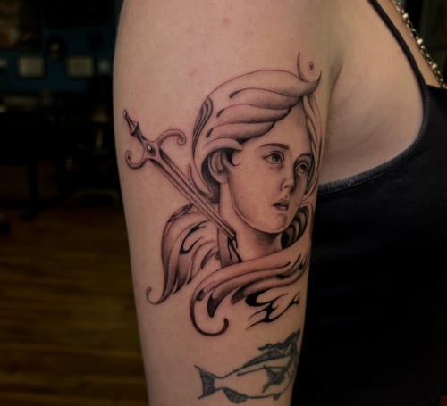 A black and grey neo-traditional tattoo on the upper arm depicting a woman's face with flowing hair and a sword behind her — by velvet devil, los angeles