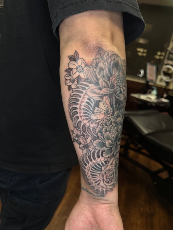 A black and grey floral arrangement tattoo on the forearm featuring detailed flowers and leaves — by Sandra Price, ventura, ca