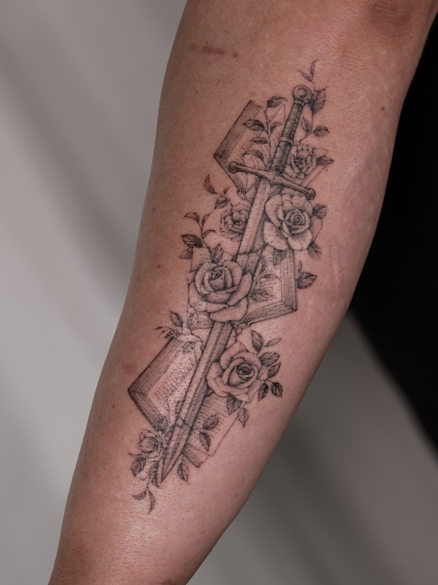 A delicate black and grey tattoo of a sword intertwined with roses and a book on the forearm, featuring fine line details and ornate design — by Melanie Benoit, los angeles