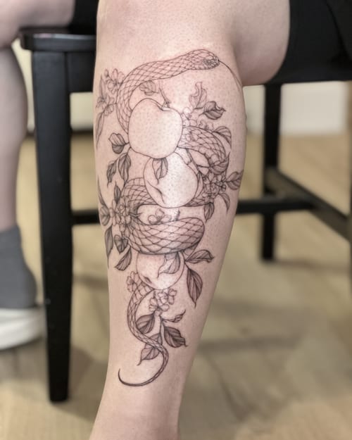 A delicate black and grey tattoo of a snake intertwined with apples and flowers on the calf, done in a fine line style — by Melanie Benoit, los angeles