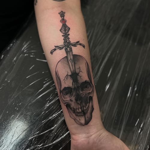 A black and grey skull with a sword on the forearm in blackwork style — by Maddie Ramirez, los angeles