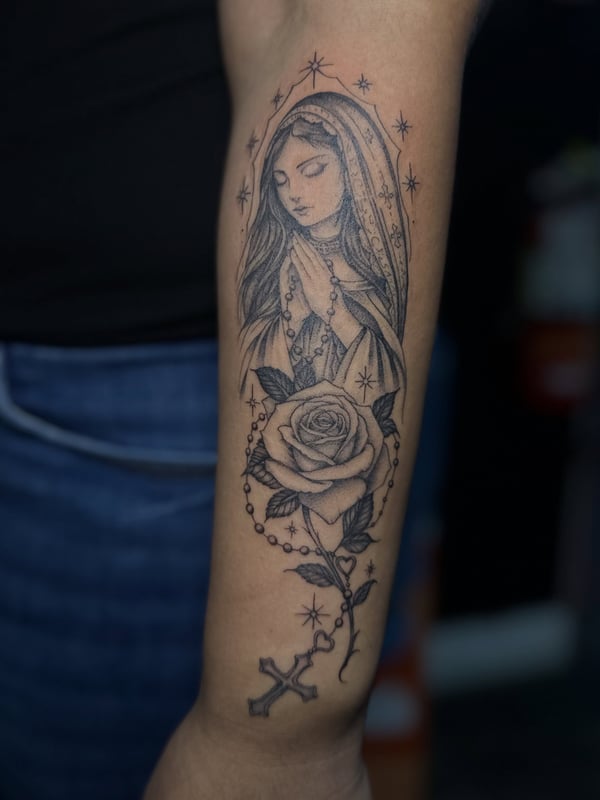 A black and grey tattoo of the Virgin Mary with a rose and rosary on the forearm, featuring fine line details and a spiritual theme — by Maddie Ramirez, palm desert, ca