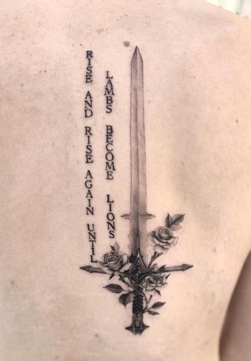 A black and grey tattoo on the back featuring a detailed sword with roses entwined around it, accompanied by a vertical quote — by otaybeatdown, los angeles