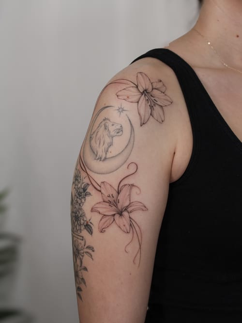 A delicate fine line tattoo of a lion and flowers with a crescent moon on the upper arm, done in black and grey — by Melanie Benoit, los angeles