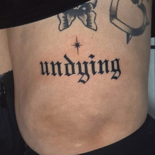 A bold black script tattoo of the word 'undying' on the rib, featuring a star above the lettering — by Miguel ANGELO, los angeles