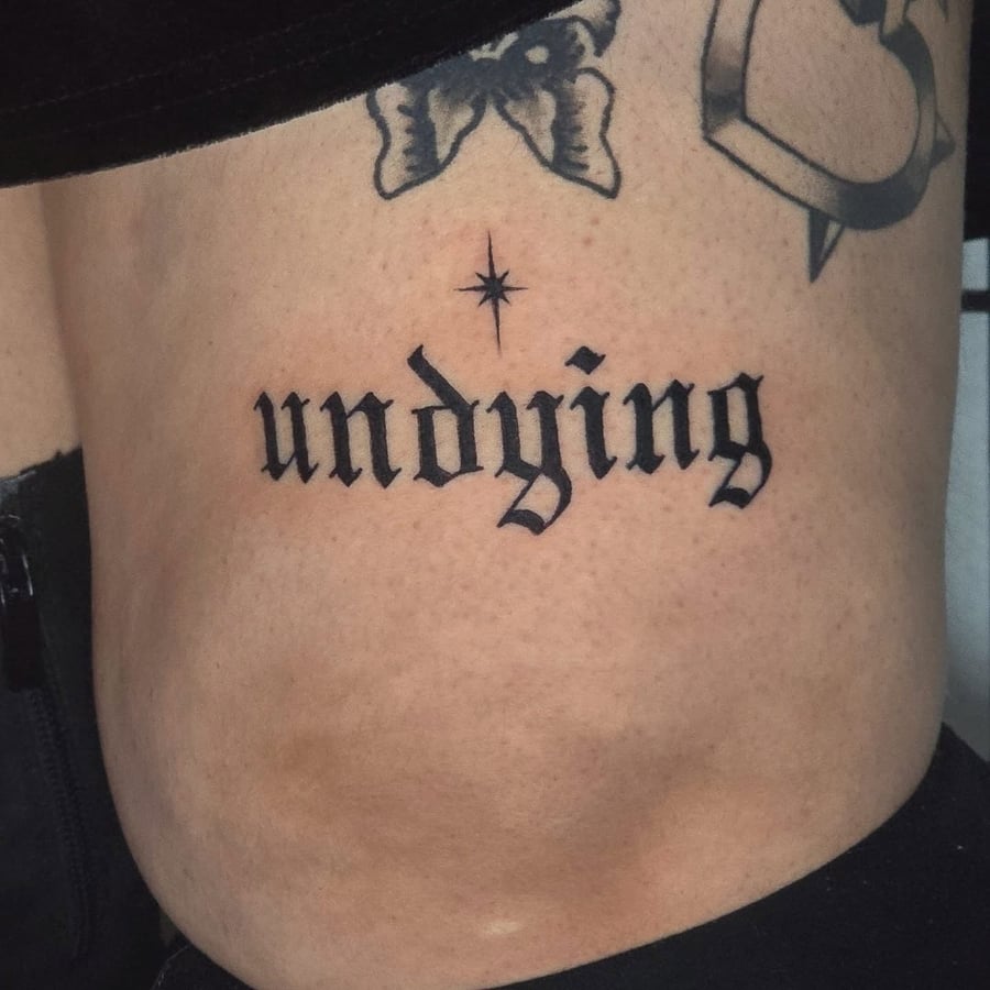 A bold black script tattoo of the word 'undying' on the rib, featuring a star above the lettering — by Miguel ANGELO, los angeles