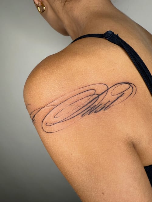 A delicate script lettering tattoo on the shoulder in black ink with fine line details — by Level tattoo, los angeles