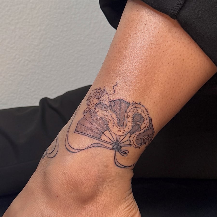 A delicate black and grey fine line tattoo of a dragon with a fan on the ankle, featuring Japanese style elements — by NJ TATTOOZ, los angeles