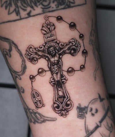 A black and grey tattoo of a crucifix with Jesus Christ and a rosary on the forearm, featuring ornate detailing — by Miguel ANGELO, los angeles
