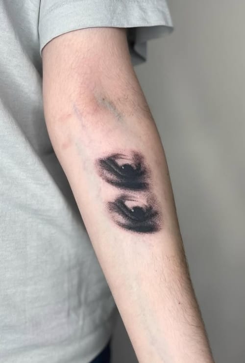 A black and grey abstract depiction of two eyes on the forearm, featuring bold blackwork with a smudged effect — by Level tattoo, los angeles